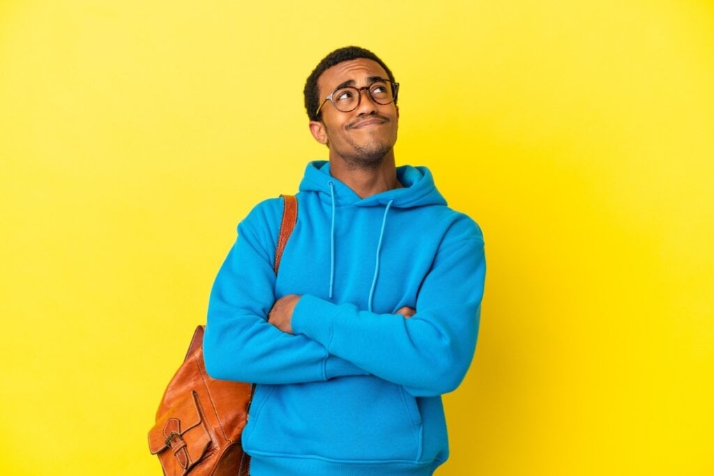 Homem vestindo um moletom azul, com uma mochila marrom nas costas, com os braços cruzados e olhando para cima pensando