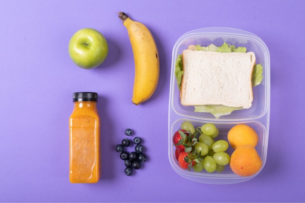 Mesa roxa com um recipiente com sanduíche natural e frutas ao lado de uma garrafa com suco laranja, banana, maçã e mirtilo