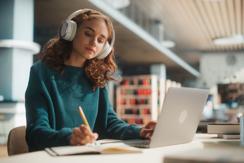 Mulher usando fones de ouvido e casaco escrevendo em caderno, que está ao lado de notebook aberto