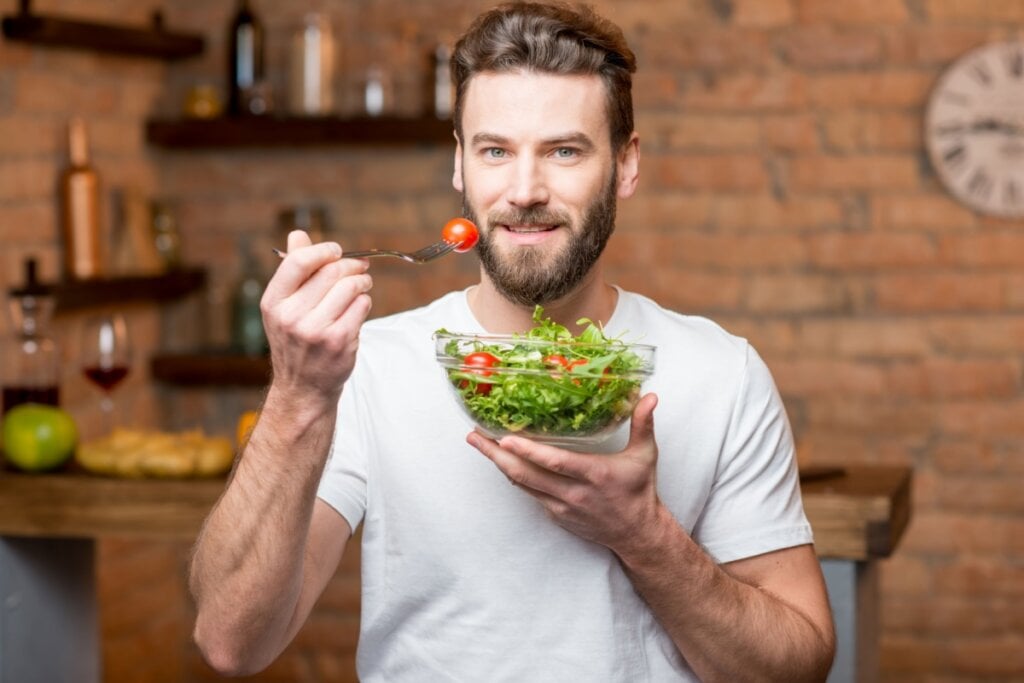 homem de camiseta branca segurando pote de salada e levando garfo com tomate à boca