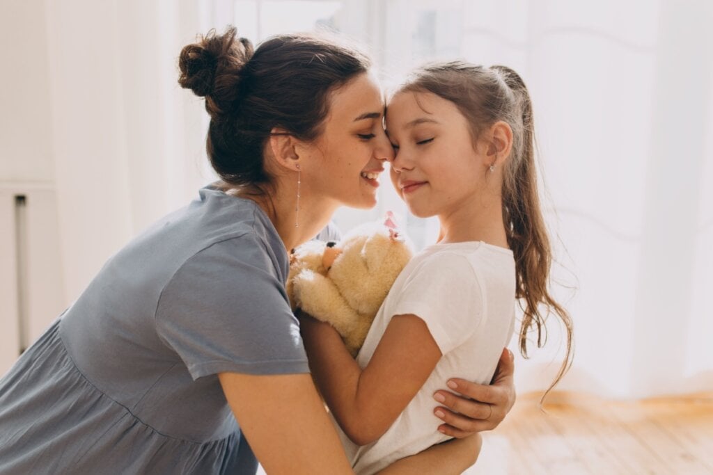 mãe abaixada abraçando menina segurando um ursinho