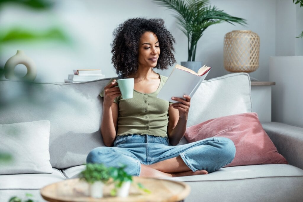 Mulher jovem negra lendo um livro sentada no sofá com uma caneca de chá na mão