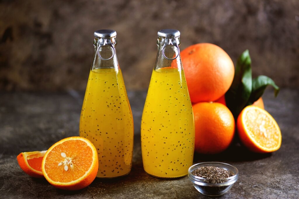 Garrafinhas de vidro contendo suco de laranja com sementes de chia, acompanhadas por laranjas frescas cortadas e um recipiente com sementes de chia