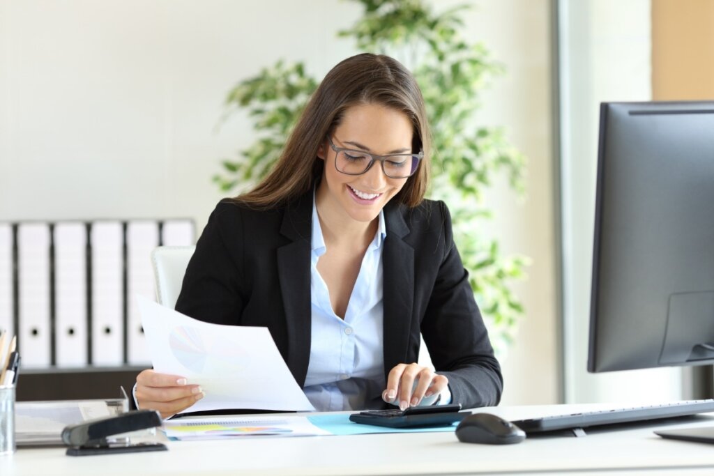 Mulher sentada, usando um terno preto e camisa social azul, sorrindo e lendo um papel em frente a um computador