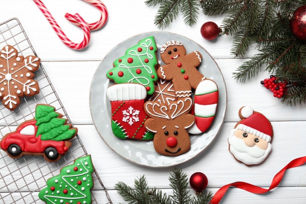 Biscoitos natalinos decorados com glacê em prato branco; ao redor, outros biscoitos e parte de árvore de Natal