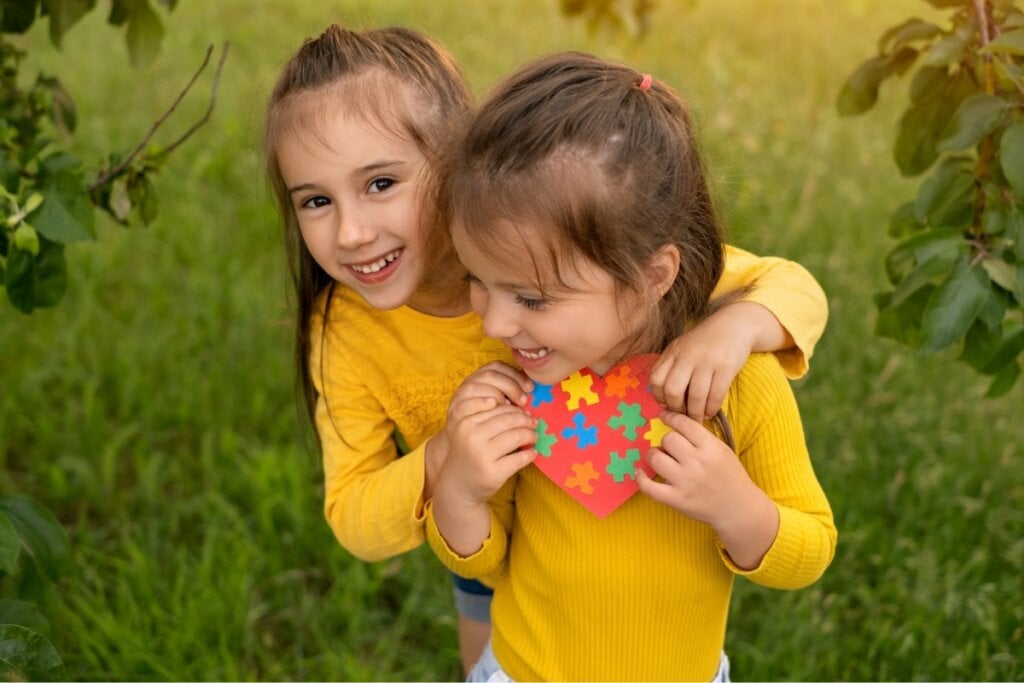 Duas meninas abraçadas, usando blusas amarelas e segurando um coração vermelho com peças de um quebra-cabeça colorido