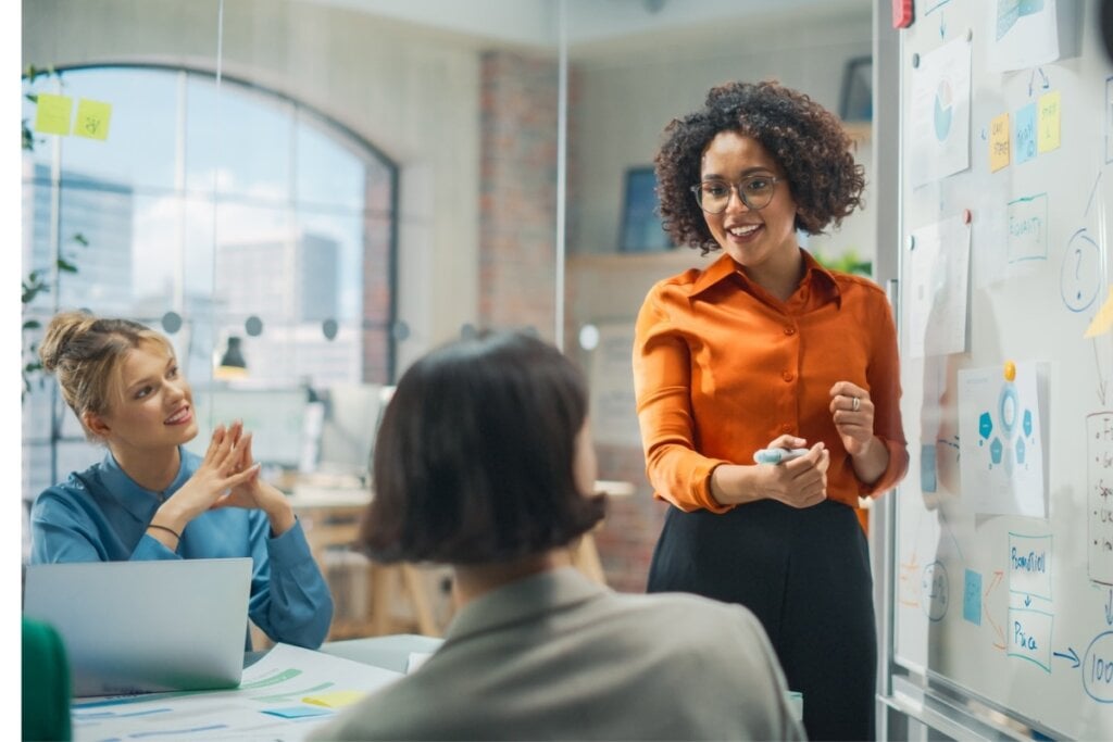 Mulher em pé escrevendo em uma lousa e conversando com duas mulheres sentadas