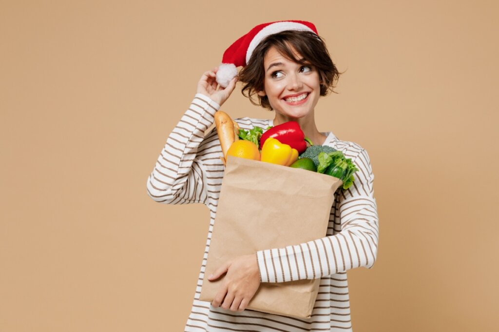 Mulher sorridente usando um gorro de Papai Noel segurando sacola de papel cheia de alimentos frescos, como pimentões, brócolis, alface, laranja