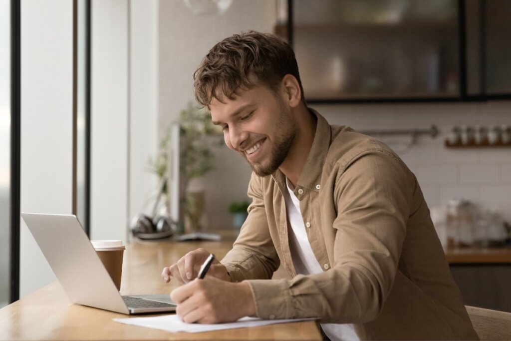 Homem jovem escrevendo em uma folha enquanto utiliza um notebook para estudos