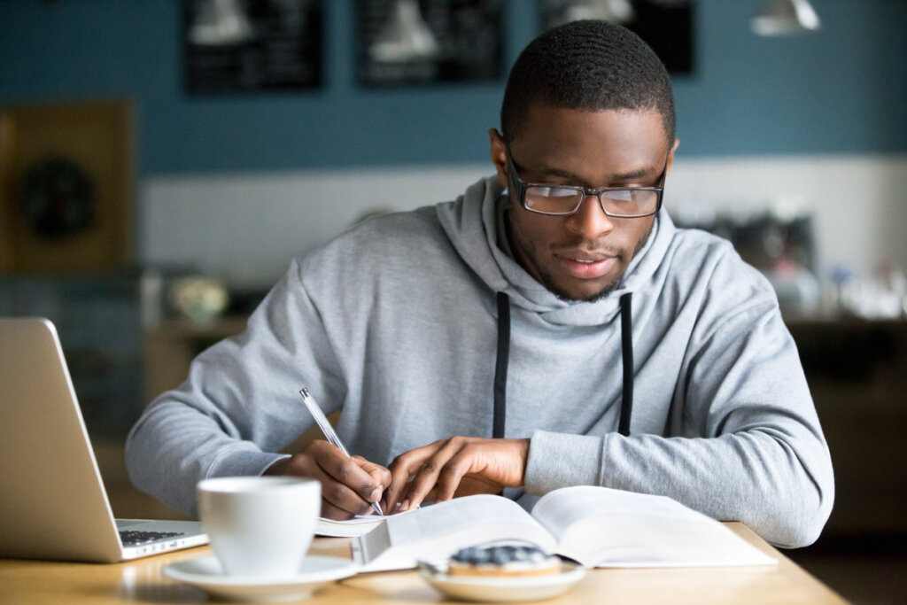 Homem negro usando casaco cinza e óculos estudando com livro; em cima da mesa, também há notebook e xícara com café