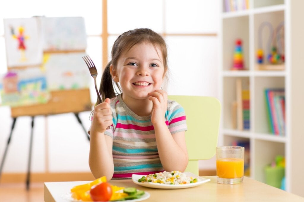 menina sentada à mesa, segurando garfo e sorrindo. na mesa há prato com comida e copo com suco