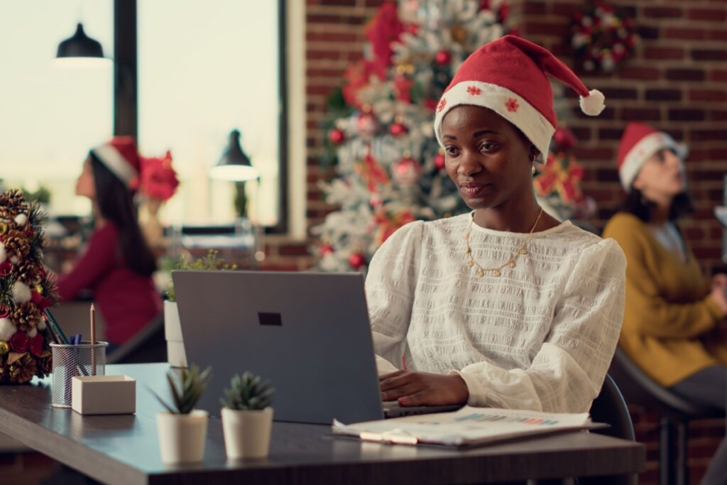 mulher com gorro de natal, sentada e mexendo em computador. árvore de natal decorada ao fundo