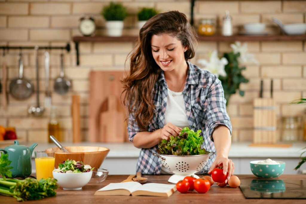 Mulher na cozinha segurando alface e tomate enquanto olha para livro em bancada