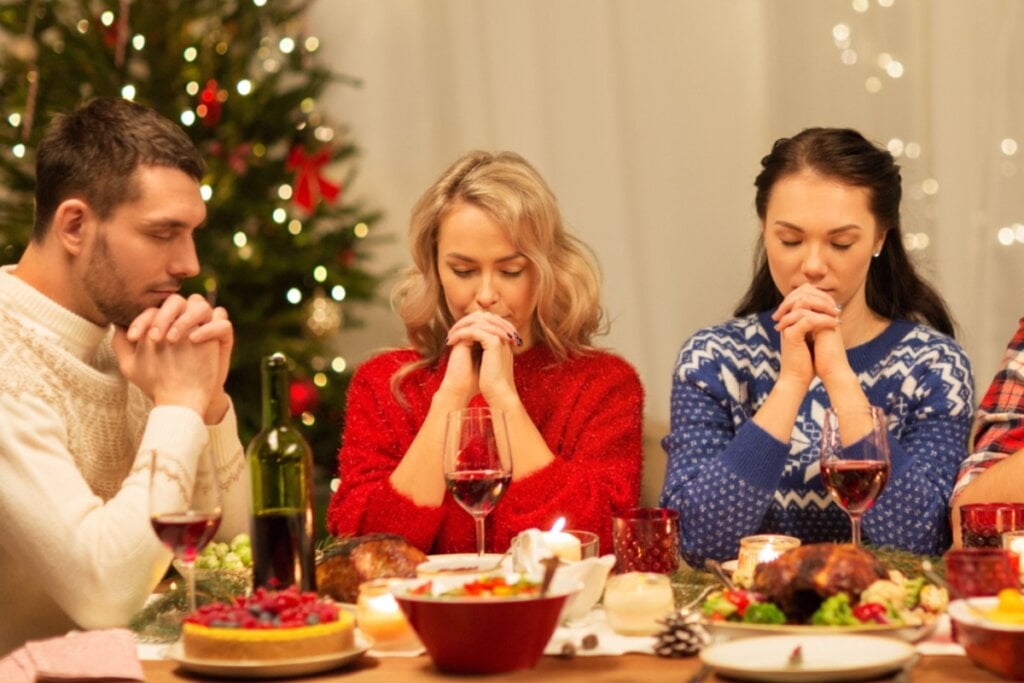 Três pessoas sentadas ao redor de uma mesa decorada para a ceia de Natal, com as mãos unidas em oração e olhos fechados