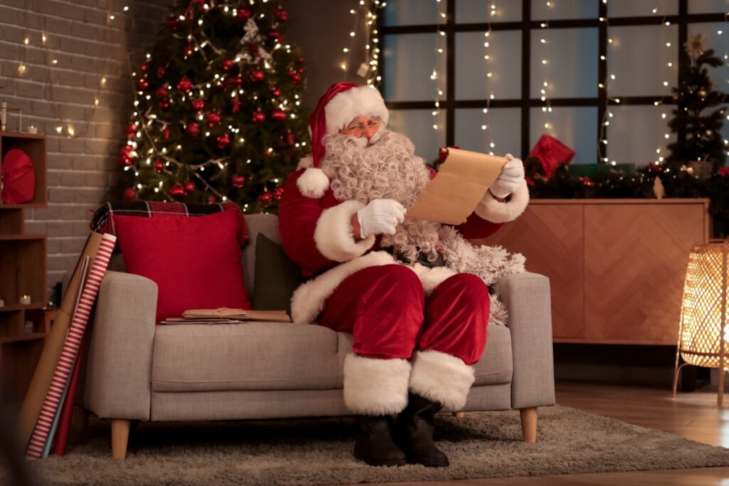 papai noel sentado no sofá e lendo papel com árvore de natal ao fundo