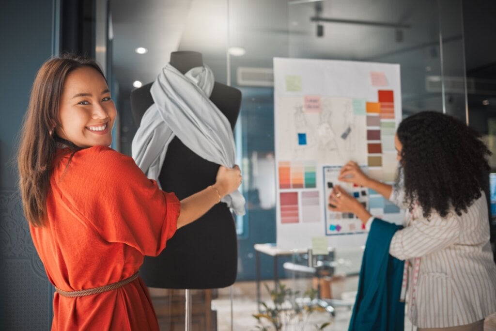 Mulher sorrindo enquanto ajusta um tecido em um manequim, enquanto outra seleciona amostras de cores e texturas em um painel