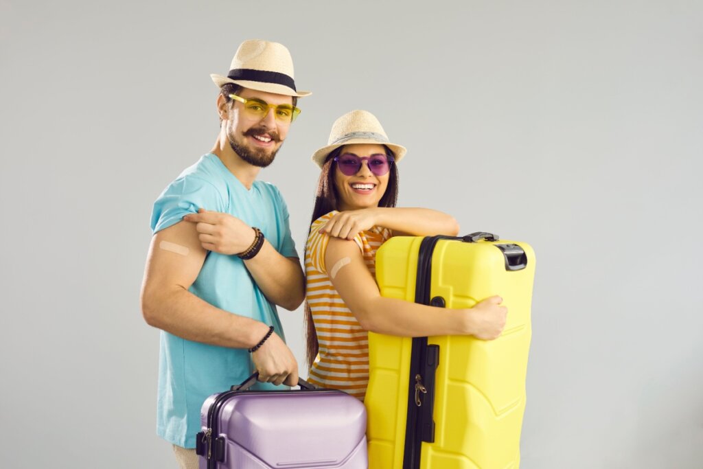 Casal sorridente com roupas de verão, chapéus e óculos de sol, mostrando adesivos no braço após vacinação e segurando malas de viagem coloridas