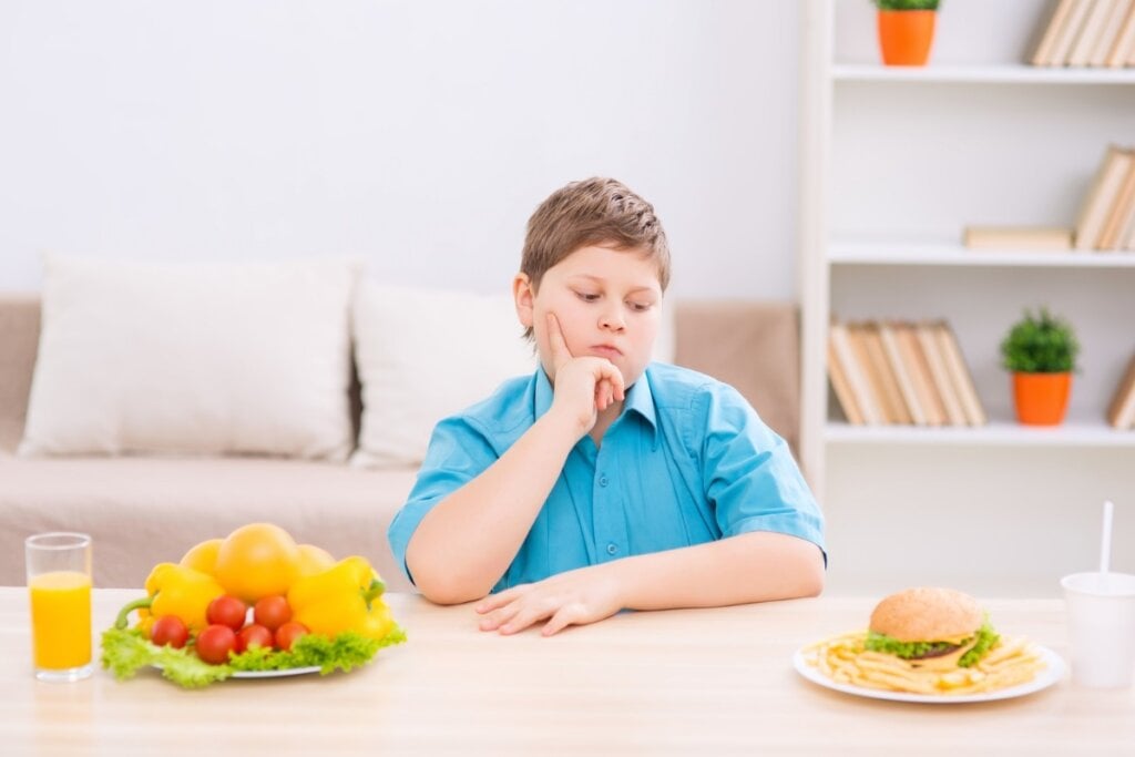 Menino usando uma camisa azul, sentado em uma mesa olhando para um prato com hambúrguer e outro prato com salada ao lado