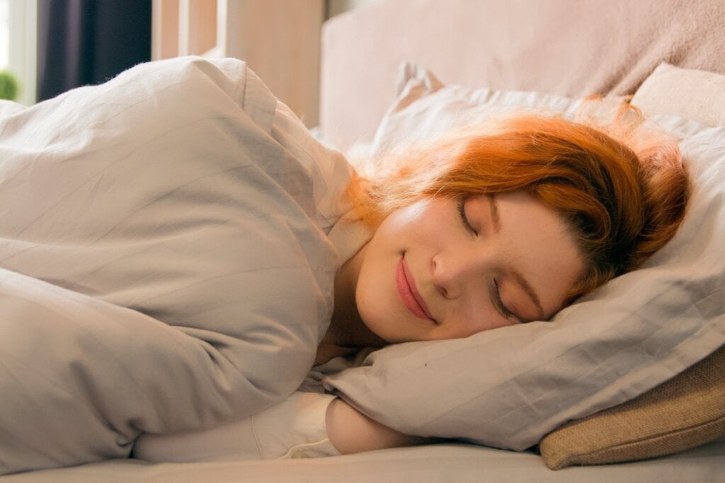 Mulher ruiva deitada em uma cama, usando um cobertor branco e com os olhos fechados e sorrindo