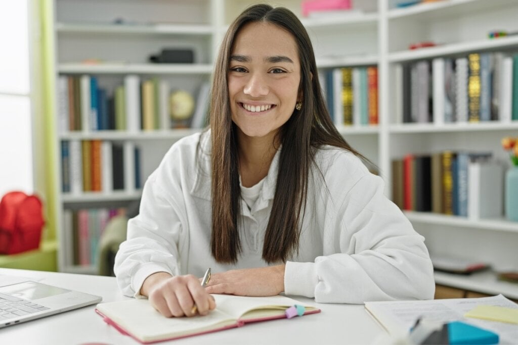 Jovem sentada em frente a uma estante com livros, segurando um lápis sobre um caderno vermelho e sorrindo