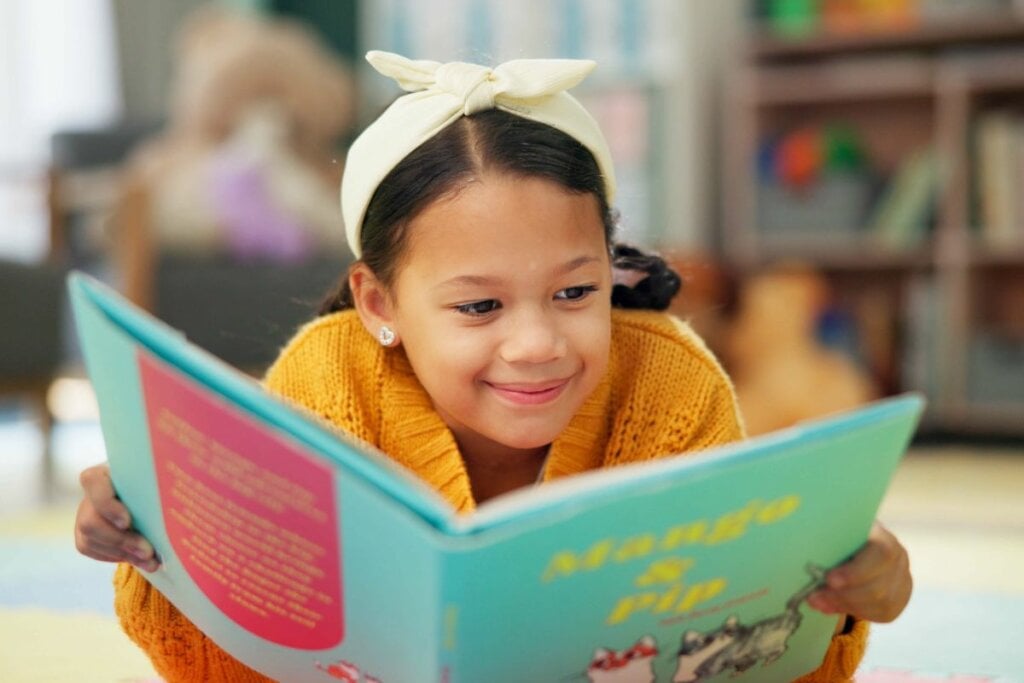 Menina sorrindo, deitada no chão e lendo um livro