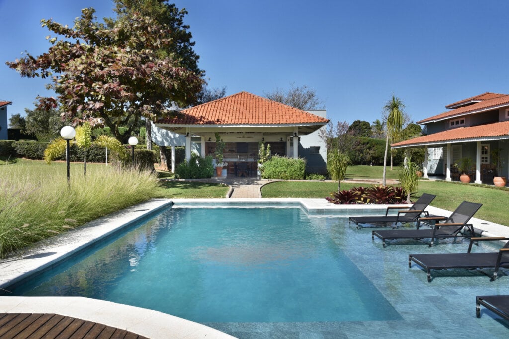 imagem de casa ao fundo, árvores em volta e piscina com espreguiçadeiras à frente