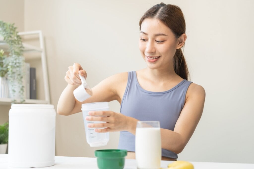 mulher de camiseta cinza colocando whey protein em colo. há banana e copo de leite na mesa