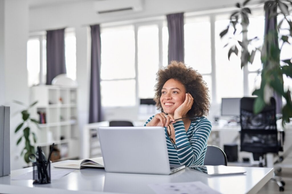 mulher sentada de frente para notebook, com mão apoiada na cabeça, olhando para cima e pensando