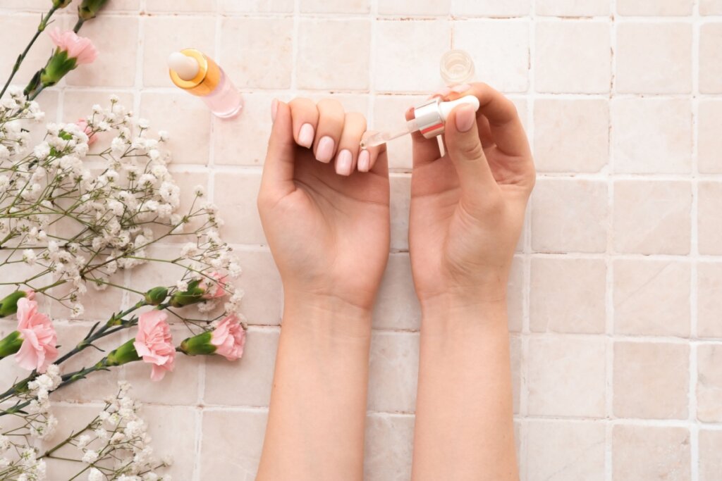 Mãos femininas aplicando óleo hidratante nas cutículas, em uma composição delicada com flores brancas e rosas sobre fundo de azulejos claros