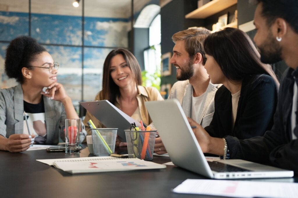 cinco pessoas reunidas em mesa de trabalho se olhando e sorrindo