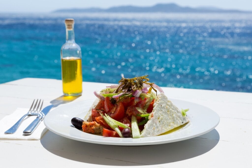 prato branco com salada grega, azeite ao lado e mar ensolarado ao funco